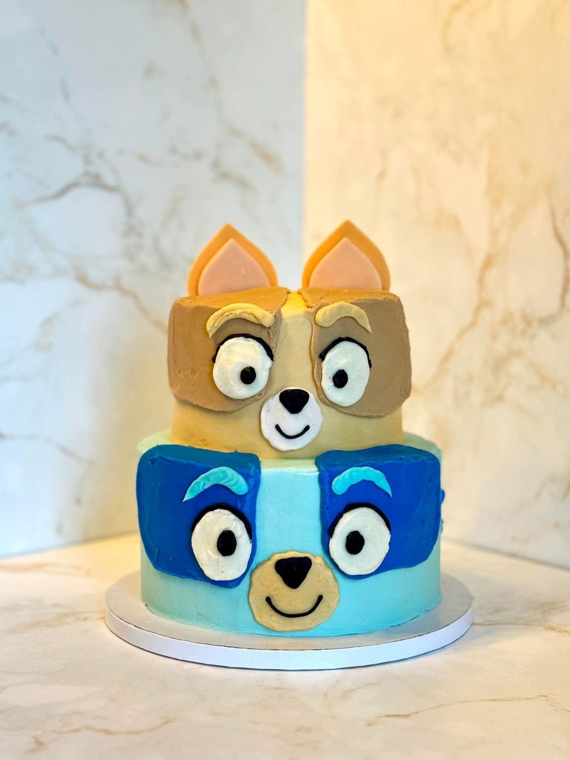 2-tier Bluey and Bingo themed children's birthday cake with character faces in blue and tan frosting