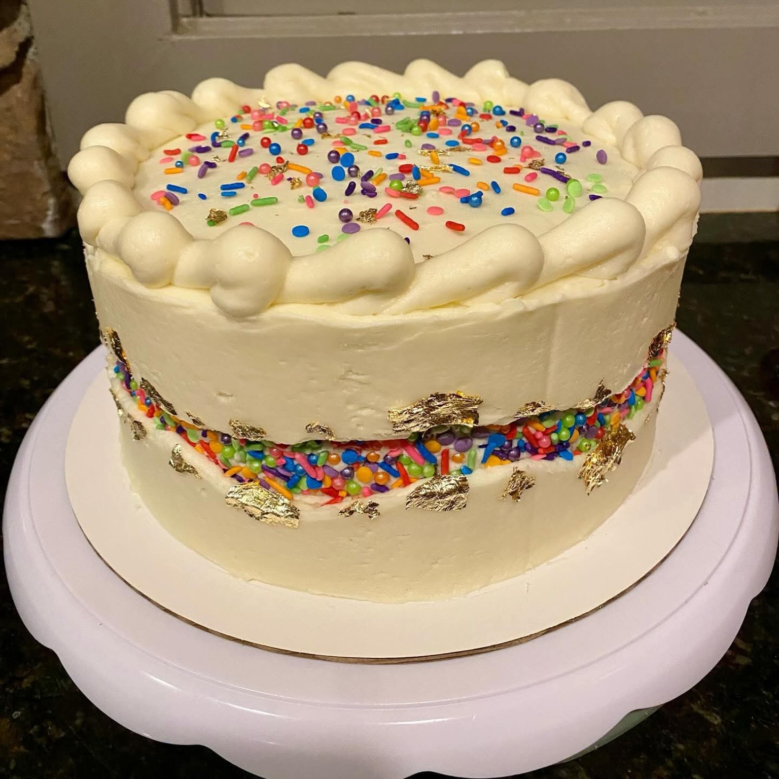 Confetti cake with white buttercream, rainbow sprinkles, and gold leaf accents on a cake stand