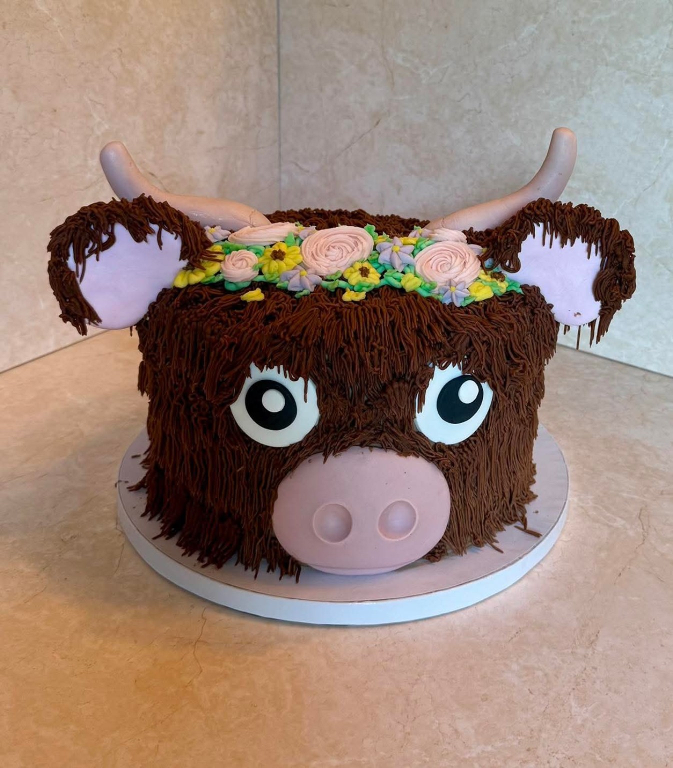 Highland cow character cake with textured brown frosting, a flower crown, and fondant facial features