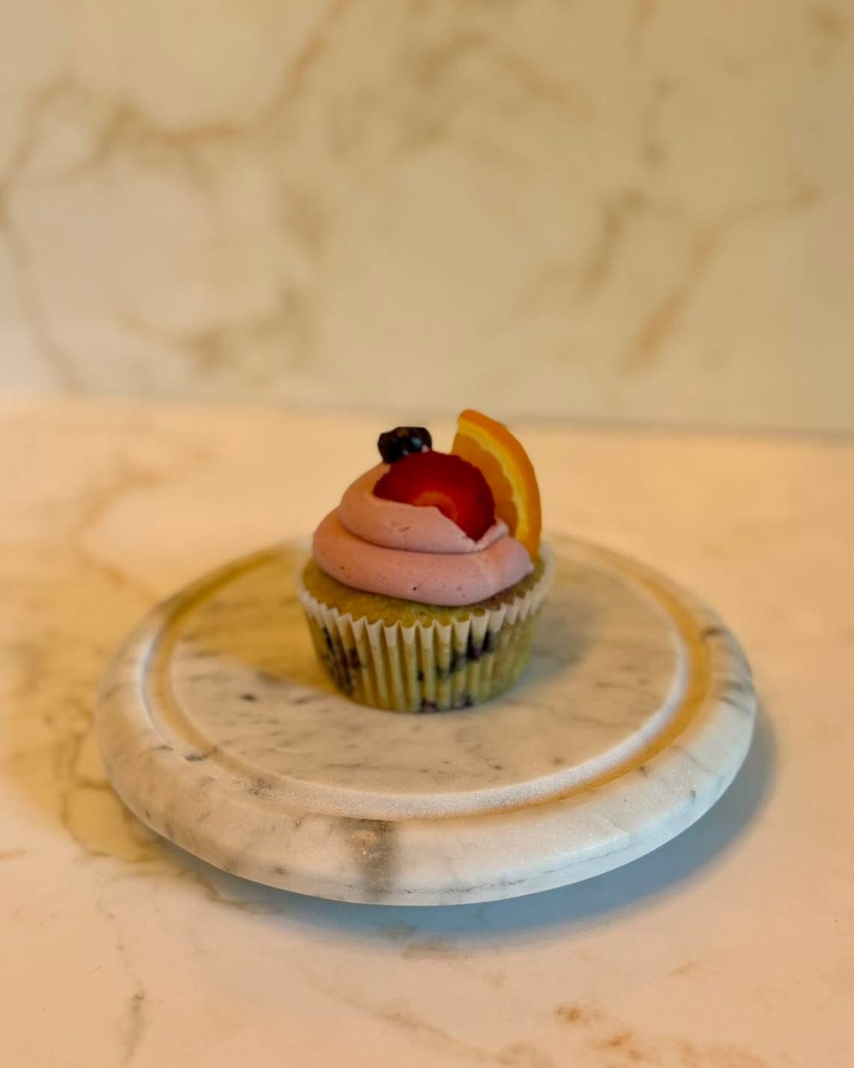 Single sangria cupcake on a marble stand with pink-purple frosting, a cherry, and an orange slice