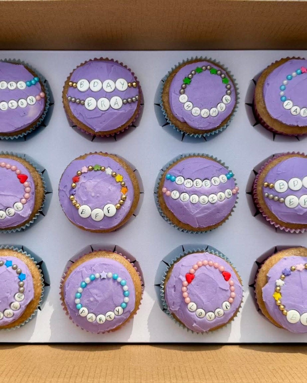 Dozen cupcakes with purple frosting and friendship bracelet bead decorations spelling out birthday messages